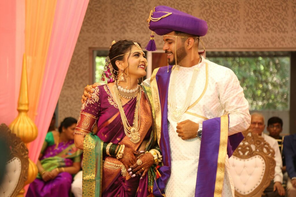 Smiling couple in traditional attire at a vibrant Indian wedding ceremony indoors.